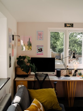A cozy and neatly organized home office setup with a large window providing natural light. The desk features a computer monitor, a stylish chair, a desk lamp, a potted plant, and various decorative items. The wall above displays several colorful art prints. A small sofa with cushions sits near the desk, contributing to a comfortable and inviting atmosphere.