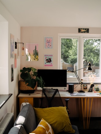 A cozy and neatly organized home office setup with a large window providing natural light. The desk features a computer monitor, a stylish chair, a desk lamp, a potted plant, and various decorative items. The wall above displays several colorful art prints. A small sofa with cushions sits near the desk, contributing to a comfortable and inviting atmosphere.