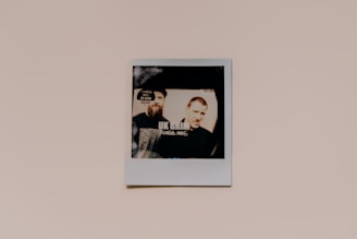 A Polaroid photo against a plain background featuring a printed image of two individuals. The photo has a vintage aesthetic with muted colors and a slightly faded appearance. The individuals appear side by side, with one wearing a hat. Some text, including 'UK Grim', is partially visible within the photo.