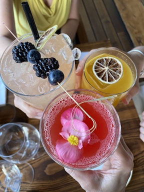 Three elegantly presented cocktails held by different hands. The first cocktail is a light pink color, garnished with blackberries and a lemon twist. The second drink is a vibrant orange hue, topped with a dried citrus slice. The third glass contains a deep pink liquid adorned with flower petals.