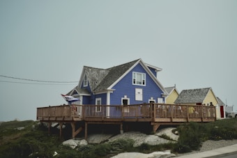 A quaint blue house with a pitched roof sits on a slightly elevated wooden deck, accompanied by smaller yellow structures nearby. The deck is adorned with a welcome flag, surrounded by rocks and patches of greenery. A cloudy or foggy sky creates a serene and calm ambiance.