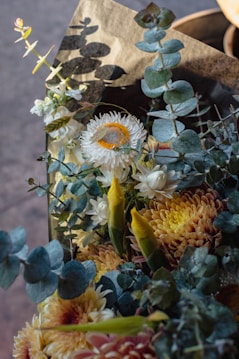 A bouquet of flowers featuring a mix of greenery and blossoms. There are sprigs of eucalyptus, strawflowers with a bright yellow center, and unopened flower buds. The arrangement is set against a neutral background with soft lighting, creating shadows.