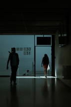 A dimly lit hallway with silhouettes of people walking towards the light. The space appears to be part of a transportation hub, possibly an airport terminal, as indicated by the presence of gate numbers B30 and B31 on the wall. The figures are carrying bags and moving towards an exit where light is streaming in.