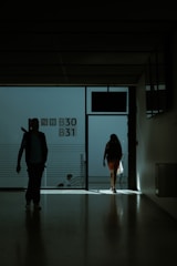 A dimly lit hallway with silhouettes of people walking towards the light. The space appears to be part of a transportation hub, possibly an airport terminal, as indicated by the presence of gate numbers B30 and B31 on the wall. The figures are carrying bags and moving towards an exit where light is streaming in.