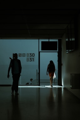 A dimly lit hallway with silhouettes of people walking towards the light. The space appears to be part of a transportation hub, possibly an airport terminal, as indicated by the presence of gate numbers B30 and B31 on the wall. The figures are carrying bags and moving towards an exit where light is streaming in.