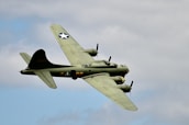 A military aircraft with four propellers is flying against a backdrop of a cloudy sky. The aircraft is olive green with distinctive markings, including a star symbol on the tail and wings, and some alphanumeric codes. The engines and propellers are visible, emphasizing its mid-flight motion.
