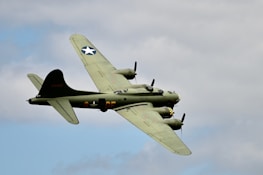 A military aircraft with four propellers is flying against a backdrop of a cloudy sky. The aircraft is olive green with distinctive markings, including a star symbol on the tail and wings, and some alphanumeric codes. The engines and propellers are visible, emphasizing its mid-flight motion.