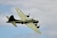 A military aircraft with four propellers is flying against a backdrop of a cloudy sky. The aircraft is olive green with distinctive markings, including a star symbol on the tail and wings, and some alphanumeric codes. The engines and propellers are visible, emphasizing its mid-flight motion.