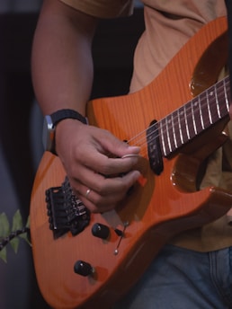 A person is playing an orange electric guitar. The focus is on the hands, with one holding a pick. The guitar has black control knobs and a wood grain finish. The person is wearing a watch on their left wrist and a casual shirt.