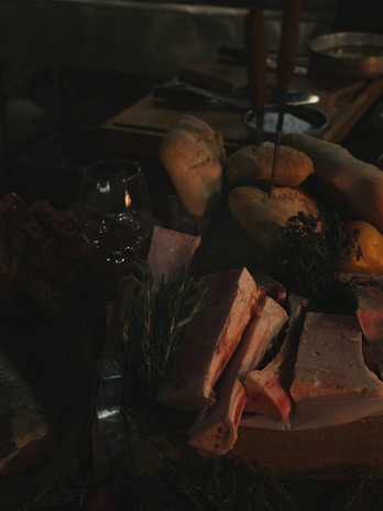 Slices of raw meat and rosemary branches are arranged on a wooden board. Surrounding the meat are loaves of bread, a glass of wine, herbs, and a lemon. The scene is dimly lit, creating a rustic and cozy atmosphere.