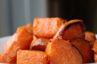 A close-up shot of a bowl filled with roasted sweet potato cubes. The pieces are glistening with a slight caramelization on their edges, indicating they are roasted and seasoned. The background is softly blurred, drawing focus to the vibrant orange of the sweet potatoes.