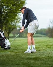 A person is preparing to hit a golf ball on a grassy course, dressed in casual sporting attire including a dark long-sleeve shirt and light shorts. The scene is set in an open area with trees and a golf bag is positioned beside the player.