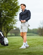 A person stands on a grassy golf course holding a golf club. They are wearing a dark long-sleeve top, light-colored shorts, and athletic shoes with socks. A golf bag is visible in the background near some trees with a cloudy sky above.
