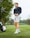 A person stands on a grassy golf course holding a golf club. They are wearing a dark long-sleeve top, light-colored shorts, and athletic shoes with socks. A golf bag is visible in the background near some trees with a cloudy sky above.