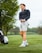 A person stands on a grassy golf course holding a golf club. They are wearing a dark long-sleeve top, light-colored shorts, and athletic shoes with socks. A golf bag is visible in the background near some trees with a cloudy sky above.
