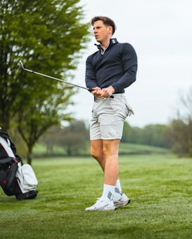 A person stands on a grassy golf course holding a golf club. They are wearing a dark long-sleeve top, light-colored shorts, and athletic shoes with socks. A golf bag is visible in the background near some trees with a cloudy sky above.