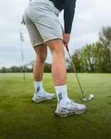 A golfer is preparing to putt on a green. The focus is on the lower body, showing white socks and athletic shoes along with a golf club and ball. The background features an open, grassy area with trees and a flag indicating the hole.