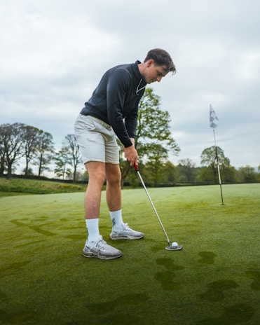 A person is putting a golf ball on a green field with a flagstick in the background, under a cloudy sky. The individual is wearing a dark long-sleeve top, light shorts, white socks, and golf shoes.