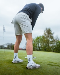 A person is focused on putting a golf ball on a green course. They are wearing a black long-sleeve top, light-colored shorts, and white athletic shoes. A golf club is held in their hands and a golf flag is visible in the background. The surroundings include lush green grass and trees under an overcast sky.