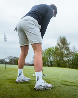 A person is focused on putting a golf ball on a green course. They are wearing a black long-sleeve top, light-colored shorts, and white athletic shoes. A golf club is held in their hands and a golf flag is visible in the background. The surroundings include lush green grass and trees under an overcast sky.