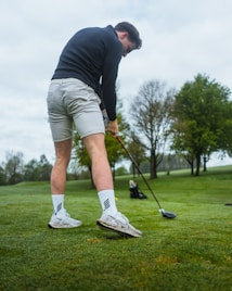 A person is preparing to take a swing on a golf course. They are wearing light-colored shorts, a dark long-sleeve top, and sports shoes. The grassy area around is well-maintained, and trees can be seen in the background.