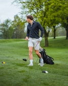 A person wearing a black jacket and light-colored shorts is standing on a lush green golf course holding a golf club. Their body is turned to the side, suggesting an action related to playing golf. A golf bag is visible in the background, and there are several trees with fresh green foliage.