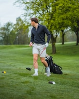 A person wearing a black jacket and light-colored shorts is standing on a lush green golf course holding a golf club. Their body is turned to the side, suggesting an action related to playing golf. A golf bag is visible in the background, and there are several trees with fresh green foliage.