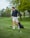 A person wearing a black jacket and light-colored shorts is standing on a lush green golf course holding a golf club. Their body is turned to the side, suggesting an action related to playing golf. A golf bag is visible in the background, and there are several trees with fresh green foliage.