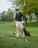 A person wearing a black jacket and light-colored shorts is standing on a lush green golf course holding a golf club. Their body is turned to the side, suggesting an action related to playing golf. A golf bag is visible in the background, and there are several trees with fresh green foliage.