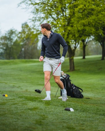A person wearing a black jacket and light-colored shorts is standing on a lush green golf course holding a golf club. Their body is turned to the side, suggesting an action related to playing golf. A golf bag is visible in the background, and there are several trees with fresh green foliage.