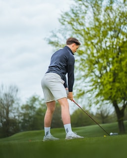 A person is preparing to hit a golf ball on a lush green course. They are wearing a dark long-sleeve shirt, light-colored shorts, and white socks with athletic shoes. The background features tall green trees and a cloudy sky.