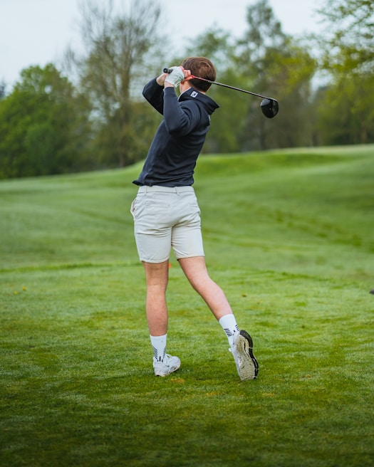 A person is swinging a golf club on a lush green course, wearing a dark long-sleeved shirt, light-colored shorts, and white sports socks with white shoes. Trees and cloudy skies are visible in the background.