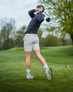 A person is playing golf on a lush green course, swinging a club. They are wearing a dark long-sleeve shirt, light shorts, and white athletic shoes. The background includes trees and a slightly overcast sky.