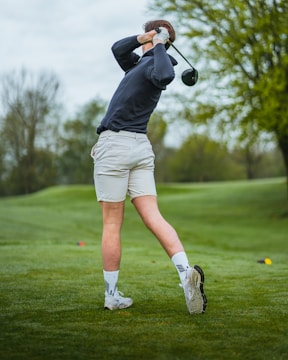 A person is playing golf on a lush green course, swinging a club. They are wearing a dark long-sleeve shirt, light shorts, and white athletic shoes. The background includes trees and a slightly overcast sky.