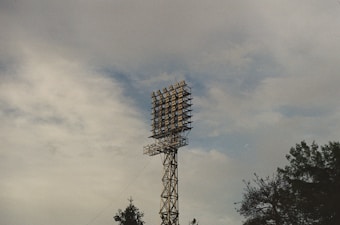 A tall metal floodlight tower stands prominently against a cloudy sky, with trees appearing at the bottom of the image. The sky is mostly overcast with patches of blue visible.