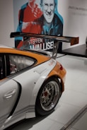 A sporty car with a large rear wing and shiny chrome wheels is displayed indoors. The rear quarter panel is prominently shown, featuring an orange and white color scheme with logos. In the background, there is a large wall mural with stylized images of people and text.