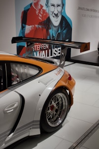 A sporty car with a large rear wing and shiny chrome wheels is displayed indoors. The rear quarter panel is prominently shown, featuring an orange and white color scheme with logos. In the background, there is a large wall mural with stylized images of people and text.