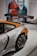 A sporty car with a large rear wing and shiny chrome wheels is displayed indoors. The rear quarter panel is prominently shown, featuring an orange and white color scheme with logos. In the background, there is a large wall mural with stylized images of people and text.