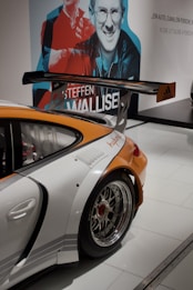 A sporty car with a large rear wing and shiny chrome wheels is displayed indoors. The rear quarter panel is prominently shown, featuring an orange and white color scheme with logos. In the background, there is a large wall mural with stylized images of people and text.
