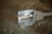 A broken streetlight fixture lies on the ground. The casing is cracked, and pieces of shattered glass are scattered around it. The streetlight is labeled with 'Pelsan' and rests on a dirt surface with some rocks and dried leaves visible.