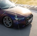 The image features a sleek, modern car with a shimmering metallic finish, displaying a play of light and shadow across its surface. The sun creates a prominent lens flare in the foreground, highlighting the car's polished appearance. A portion of the license plate is visible, as well as the car&rsquo;s stylish wheels and sharp headlights. The background shows a paved road bordered by a curb with grassy terrain beyond.