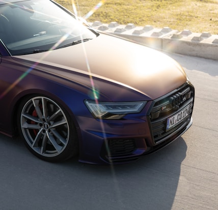 The image features a sleek, modern car with a shimmering metallic finish, displaying a play of light and shadow across its surface. The sun creates a prominent lens flare in the foreground, highlighting the car's polished appearance. A portion of the license plate is visible, as well as the car&rsquo;s stylish wheels and sharp headlights. The background shows a paved road bordered by a curb with grassy terrain beyond.