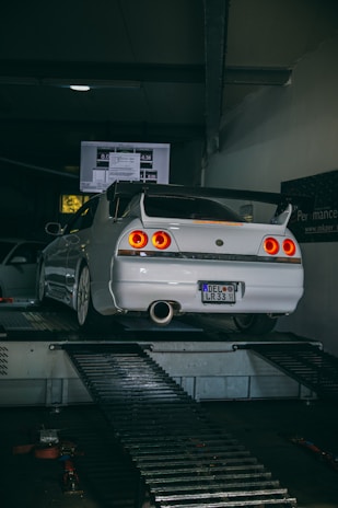 A white sports car is positioned on a dynamometer in a dimly lit garage. The car's rear lights are illuminated, and there is a screen displaying data in the background. The room is industrial in design with metal platforms and equipment surrounding the vehicle.