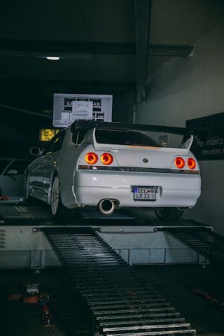A white sports car is positioned on a dynamometer in a dimly lit garage. The car's rear lights are illuminated, and there is a screen displaying data in the background. The room is industrial in design with metal platforms and equipment surrounding the vehicle.