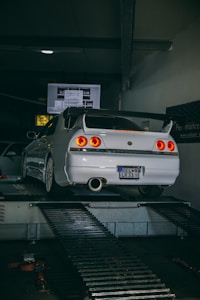 A white sports car is positioned on a dynamometer in a dimly lit garage. The car's rear lights are illuminated, and there is a screen displaying data in the background. The room is industrial in design with metal platforms and equipment surrounding the vehicle.