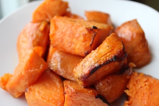 Close-up of roasted sweet potato chunks on a white plate. The pieces are golden-brown with some caramelized edges, glistening with oil or glaze.