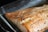 A cooked salmon fillet seasoned with pepper and various spices is placed in a glass baking dish. The salmon has a golden-brown finish, indicating it is well-cooked, with visible grill marks and a slightly flaky texture.