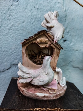 A decorative figurine of two white doves is perched on a small wooden birdhouse with the word 'LOVE' inscribed above the entry. The birdhouse has a rustic appearance with twigs inside, and the overall composition creates a peaceful and romantic scene.