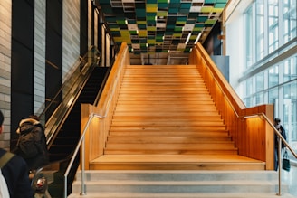 A modern indoor space featuring a wide, wooden staircase with metal railings leading upwards. To the left, an escalator ascends parallel to the stairs. The ceiling is adorned with colorful square panels, adding a contemporary feel to the space. Large windows on the right allow for ample natural light, enhancing the open, airy atmosphere. A person wearing a coat is visible near the escalator.