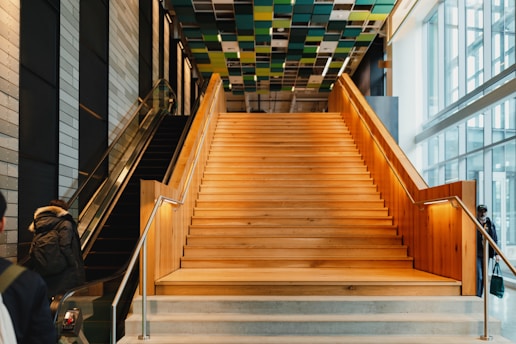 A modern indoor space featuring a wide, wooden staircase with metal railings leading upwards. To the left, an escalator ascends parallel to the stairs. The ceiling is adorned with colorful square panels, adding a contemporary feel to the space. Large windows on the right allow for ample natural light, enhancing the open, airy atmosphere. A person wearing a coat is visible near the escalator.